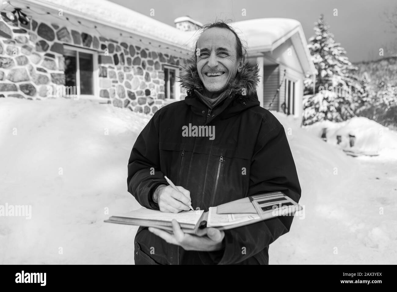 Centrale invecchiato home Inspector prendere appunti durante un'ispezione. Uomo sorridente si alza fuori nella neve con la casa sullo sfondo, in bianco e nero Foto Stock