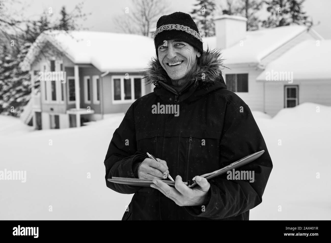Home Ispettore durante l'ispezione, uomo caucasico sorridente e tenendo un taccuino, prese appunti mentre si trova all'aperto nella neve con la casa sullo sfondo Foto Stock