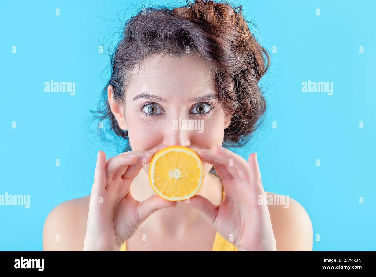 Bella brunetta in cima giallo tiene arancione davanti alla bocca su sfondo blu, concetto di bellezza Foto Stock