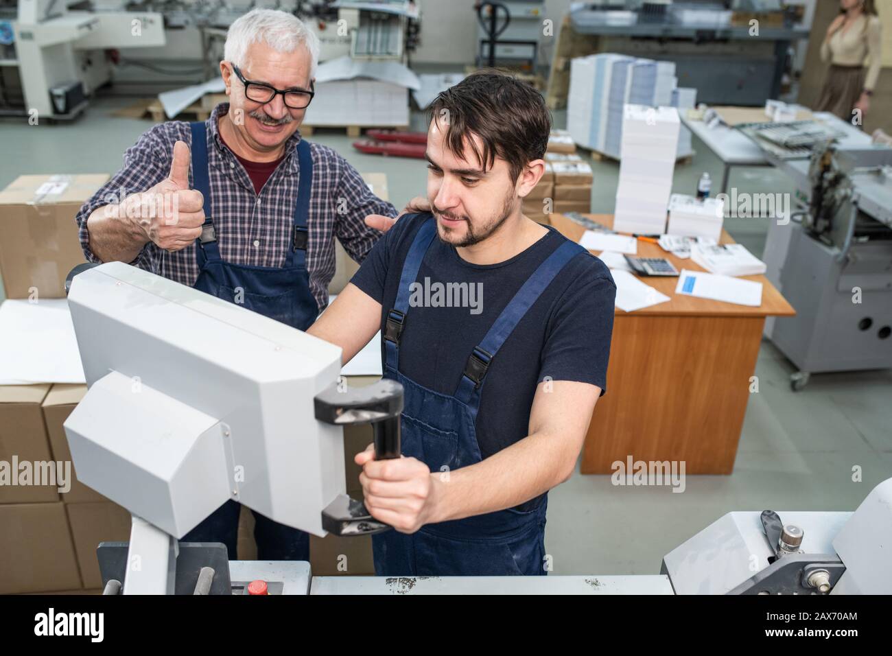 L'uomo anziano soddisfatto che mostra il pollice-in su mentre dà la valutazione alla macchina operativa intern giovane all'impianto di stampa Foto Stock