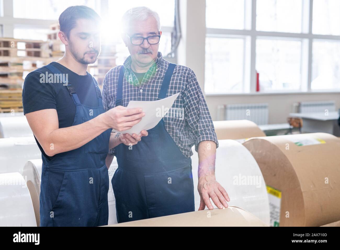 I lavoratori seri della casa di stampa nelle tute che visualizzano le note in carta mentre contano i rotoli di carta al magazzino Foto Stock
