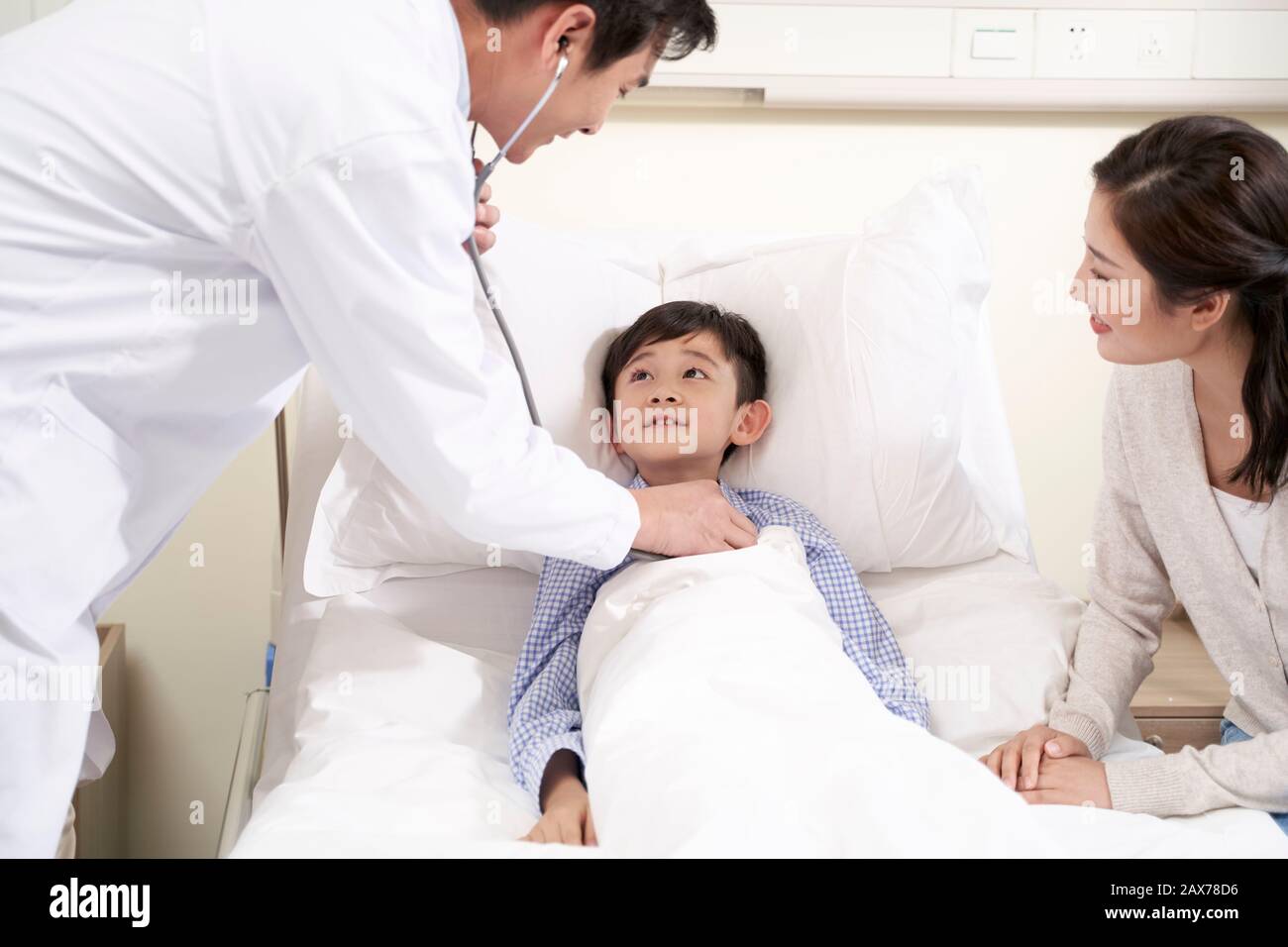 Cinque anni ragazzo asiatico che giace nel letto di ospedale accompagnati dalla madre e di essere esaminati dal pediatra utilizzando uno stetoscopio Foto Stock