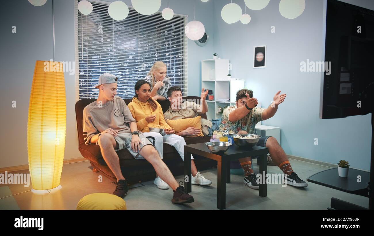 Ragazzi e ragazze che chiacchierano a casa e discutono di azione in TV mentre mangiano spuntini. Ci sono grattacieli sullo sfondo. Foto Stock