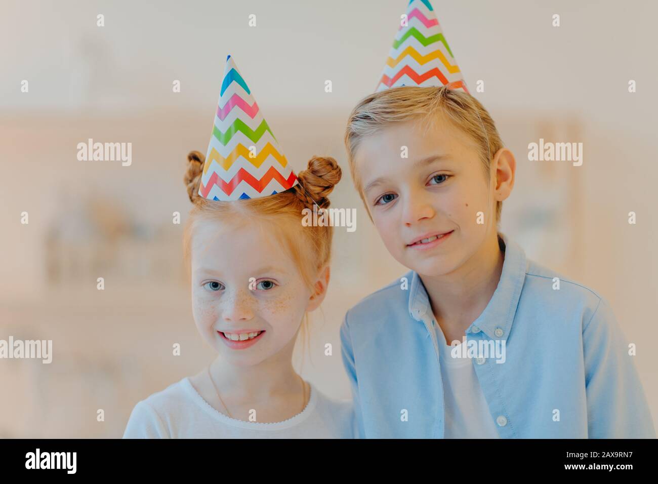 Ritratto della ragazza dello zenzero felice e il suo broghter si levano in piedi vicino a vicenda, indossano i cappelli della festa, vengono sul compleanno degli amici, osservano gladfully alla macchina fotografica, posano Foto Stock