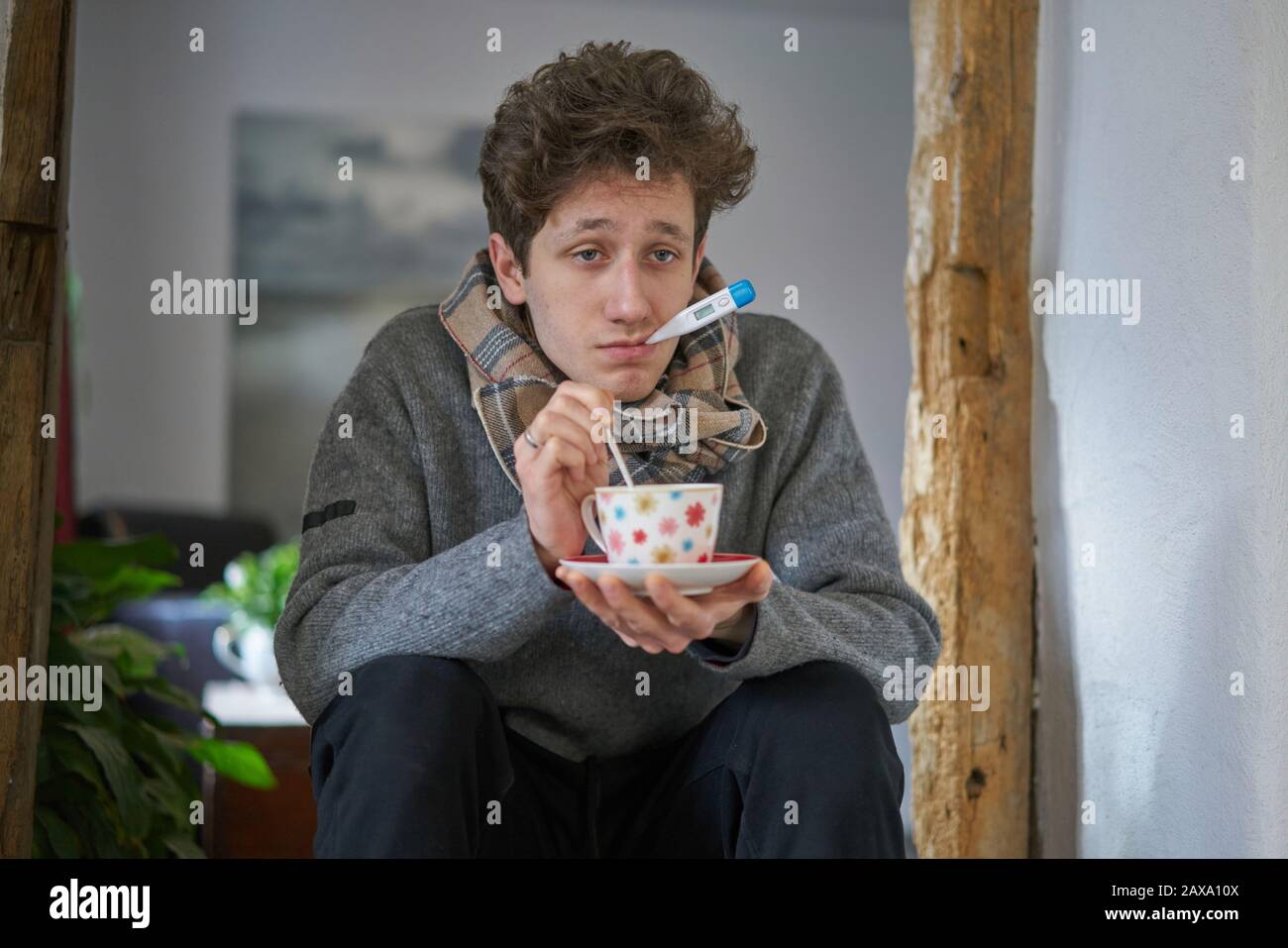 Un giovane malato con un freddo seduto in casa ha un termometro in bocca e sta tenendo una tazza in mano Foto Stock