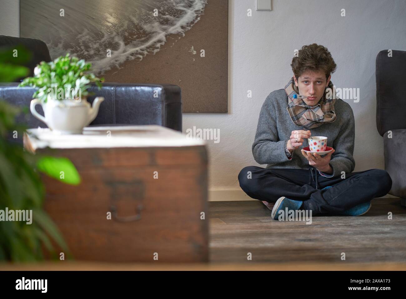 Giovane bell'uomo con un freddo seduto sul pavimento nel suo appartamento con un termometro in bocca e una tazza di tè nelle sue mani, guardando depresso Foto Stock