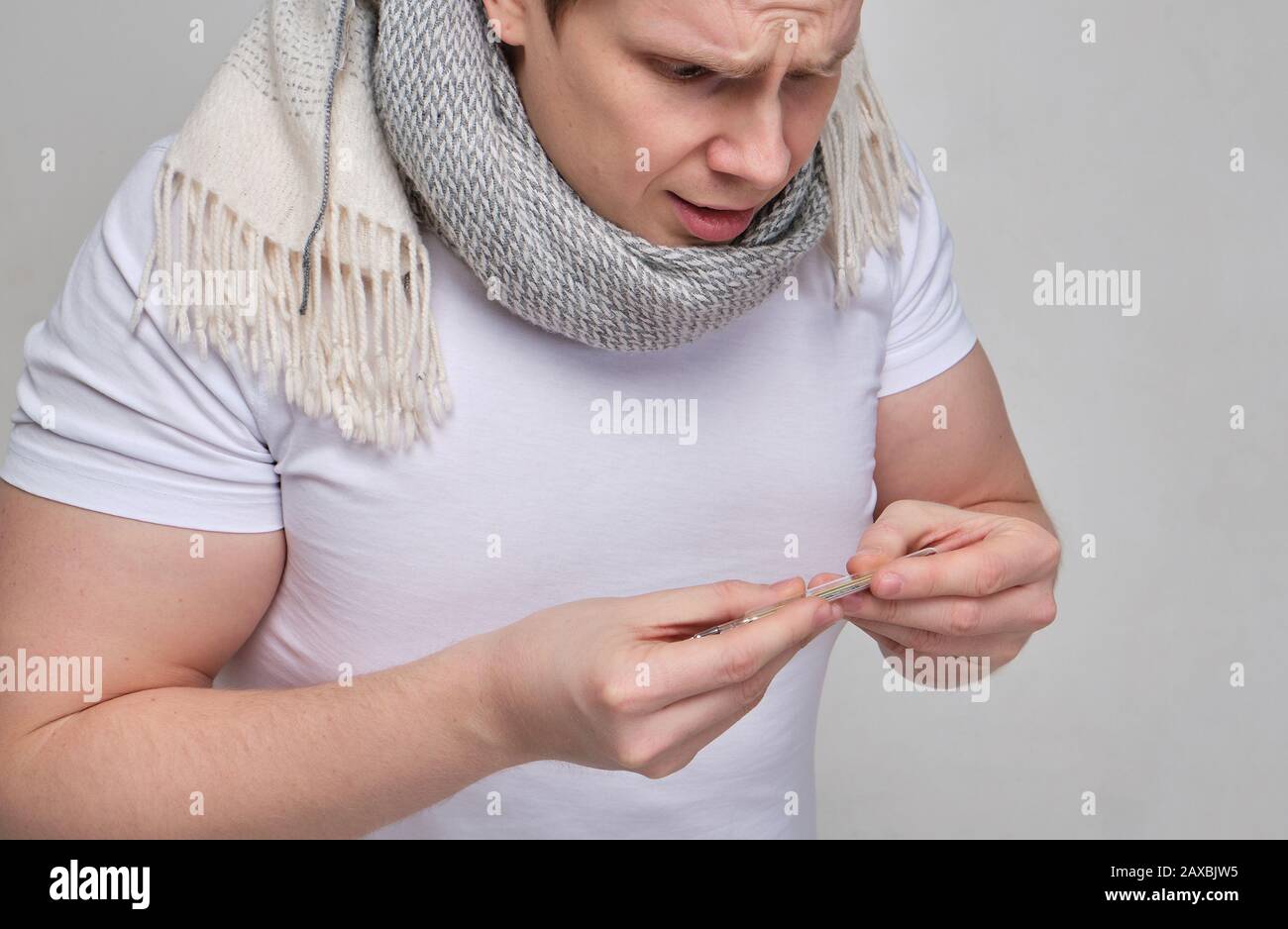 Un uomo in una maglietta bianca con una sciarpa guarda il termometro a sorpresa. Il concetto di temperatura corporea ridotta. Primo piano. Foto Stock