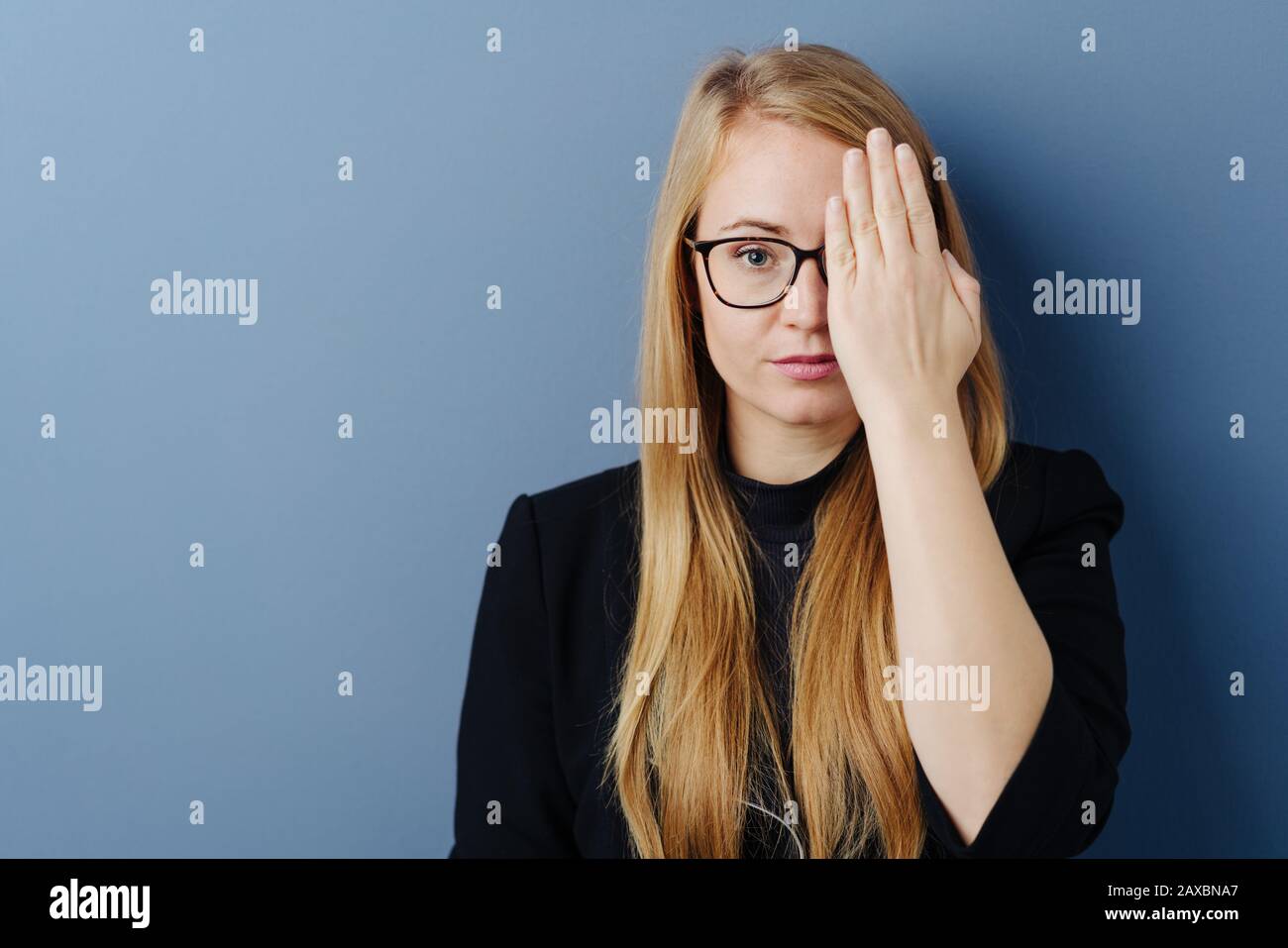 Giovane donna con lunghi capelli biondi in occhiali che la controllano la  vista, coprendo un occhio con la mano e guardando verso la fotocamera.  Primo piano verticale contro b Foto stock -