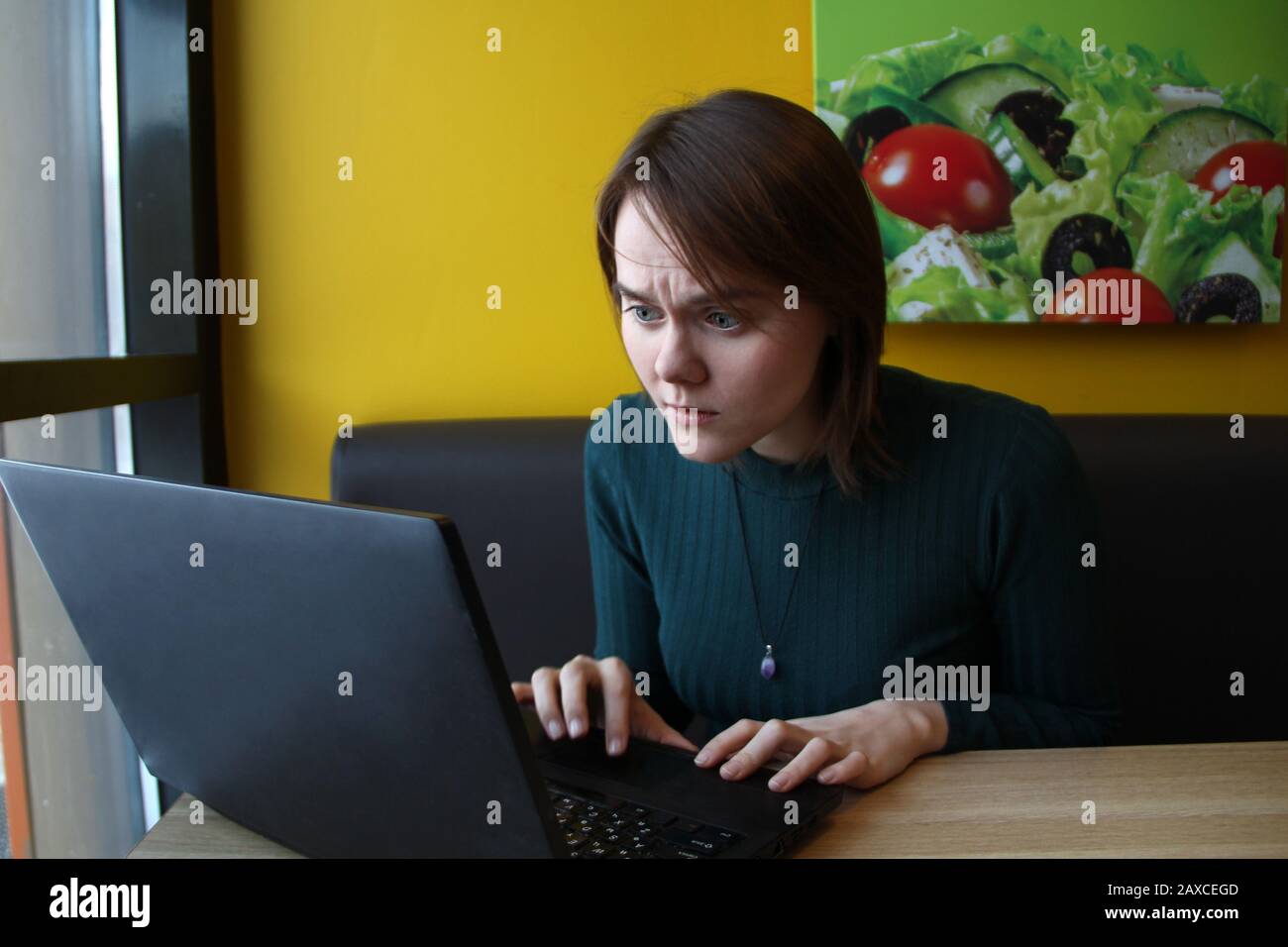 Ragazza con un'espressione di tensione perplessa si siede seduta lavorando ad un portatile ad un tavolo in un caffè su un divano marrone contro un muro giallo. Foto Stock