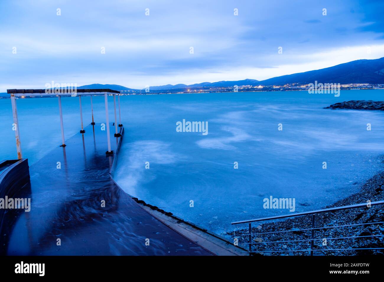 Spiaggia di Gelendzhik la sera in una tempesta. Le onde ad alta velocità si trasformano in una nebbia blu. Toni blu, inverno. Il molo esce al mare. Spiaggia di ciottoli. Foto Stock