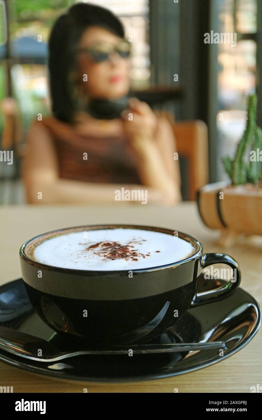 Closeup cappuccino in una tazza nera con donna sfocata bere caffè sullo sfondo Foto Stock