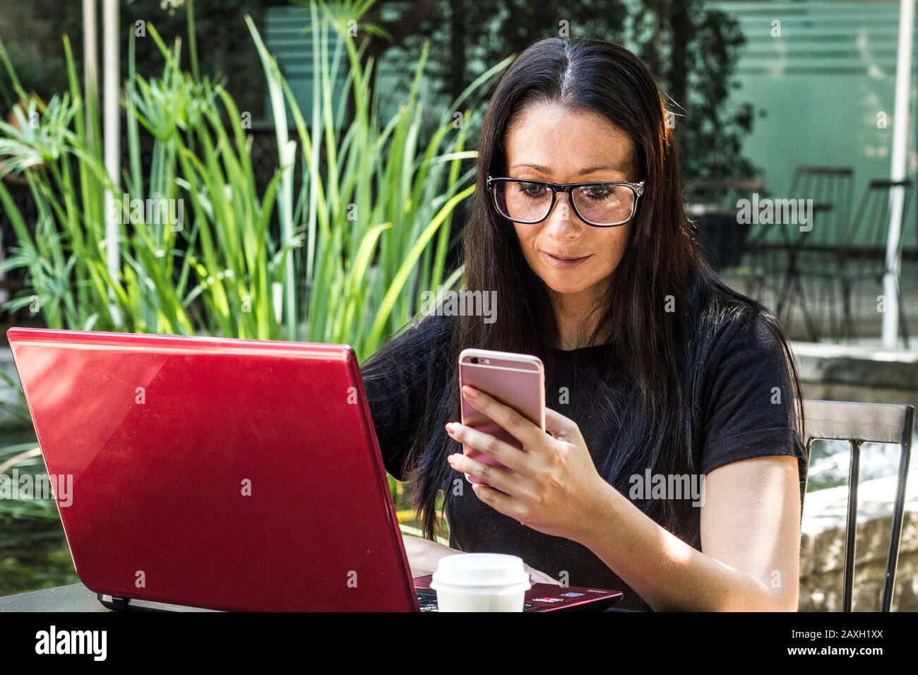 Donna d'affari con occhiali che texting un messaggio sul cellulare mentre controlla il suo laptop rosso Foto Stock
