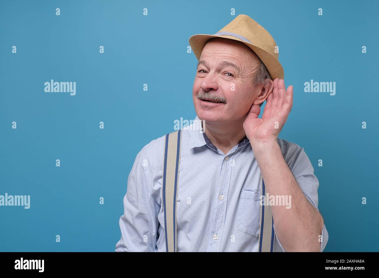 Curioso uomo anziano in estate cappello ascolto di pettegolezzi essere interessati alle ultime notizie. Studio girato su parete blu. Foto Stock