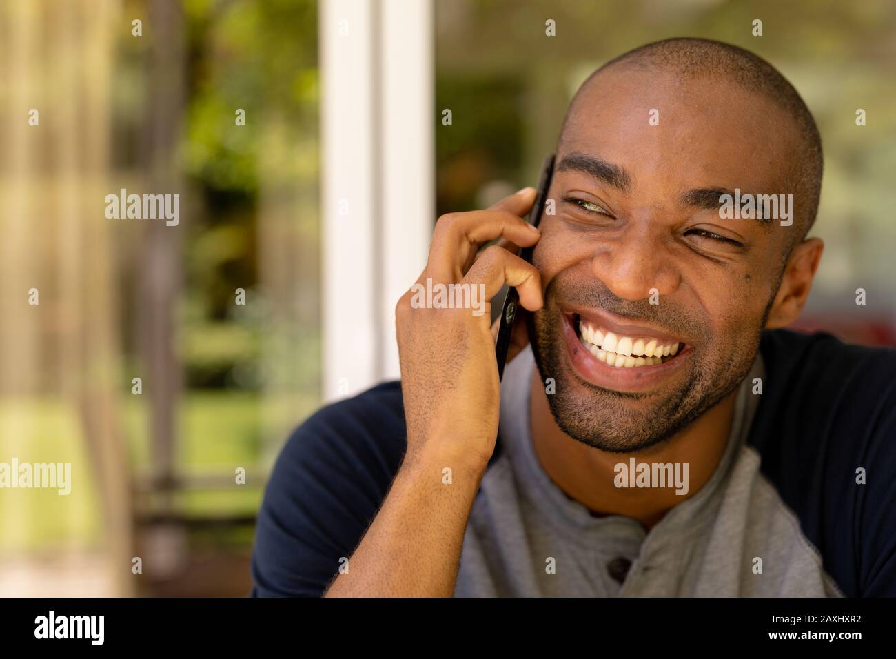 Ritratto di un uomo africano americano con t shirt grigia seduta nel patio, parlando sul suo cellulare e ridere durante una giornata di sole Foto Stock