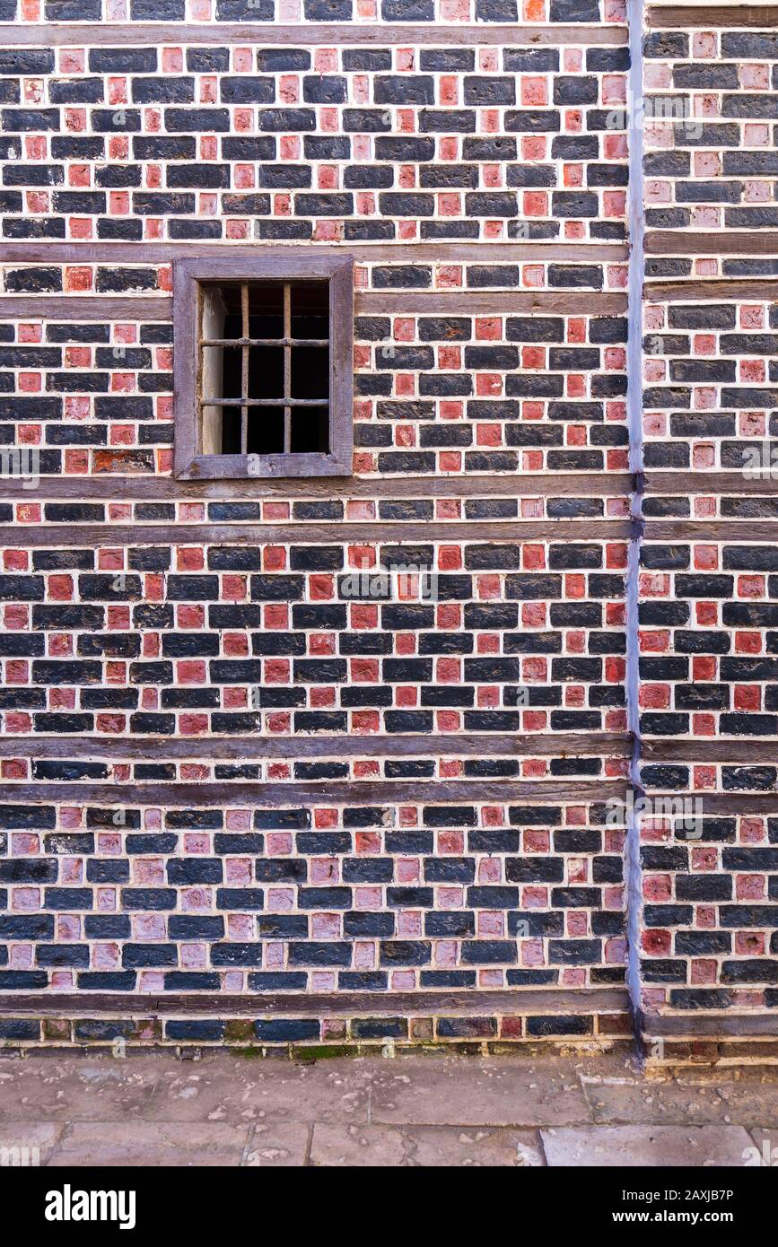 Muro con mattoni neri e rossi con cucitura bianca e piccola finestra con barre di metallo su strada acciottolata Foto Stock