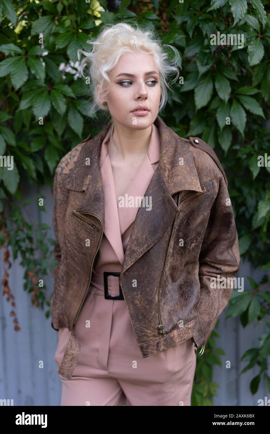 Ragazza bionda sicura in giacca vintage in pelle con le mani in tasche davanti alle piante da arrampicata. Foto Stock Ragazza bionda sicura in giacca vintage in pelle con le mani in tasche davanti alle piante da arrampicata. Foto Stock