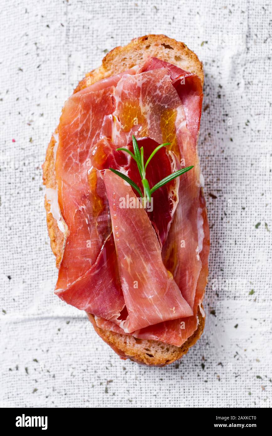 vista ad alto angolo del tomaquet spagnolo pa amb, una fetta di pane con pomodoro, sormontata da prosciutto serrano su uno sfondo di tessuto bianco testurizzato Foto Stock