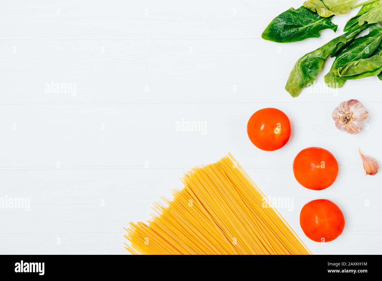 Spaghetti di grano duro crudo accanto a aglio fresco, pomodori e foglie di spinaci su sfondo bianco con spazio copia. Foto Stock