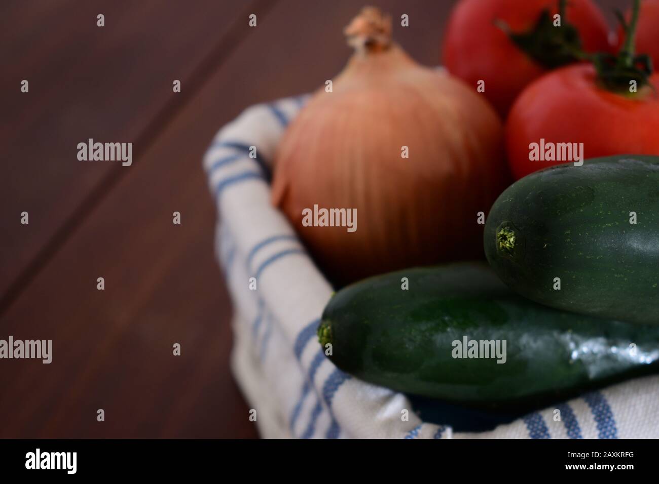 Cetrioli all'interno di un cesto con pomodori sfocati e una cipolla sullo sfondo su un tavolo di legno marrone scuro Foto Stock