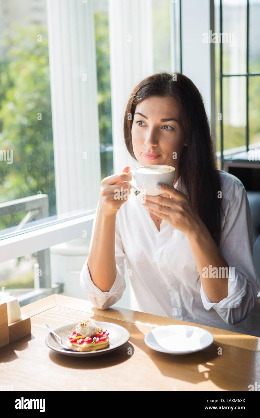 Bella sorridente giovane donna in cafe vicino alla finestra con caffè e deliziosi dolci. Foto Stock