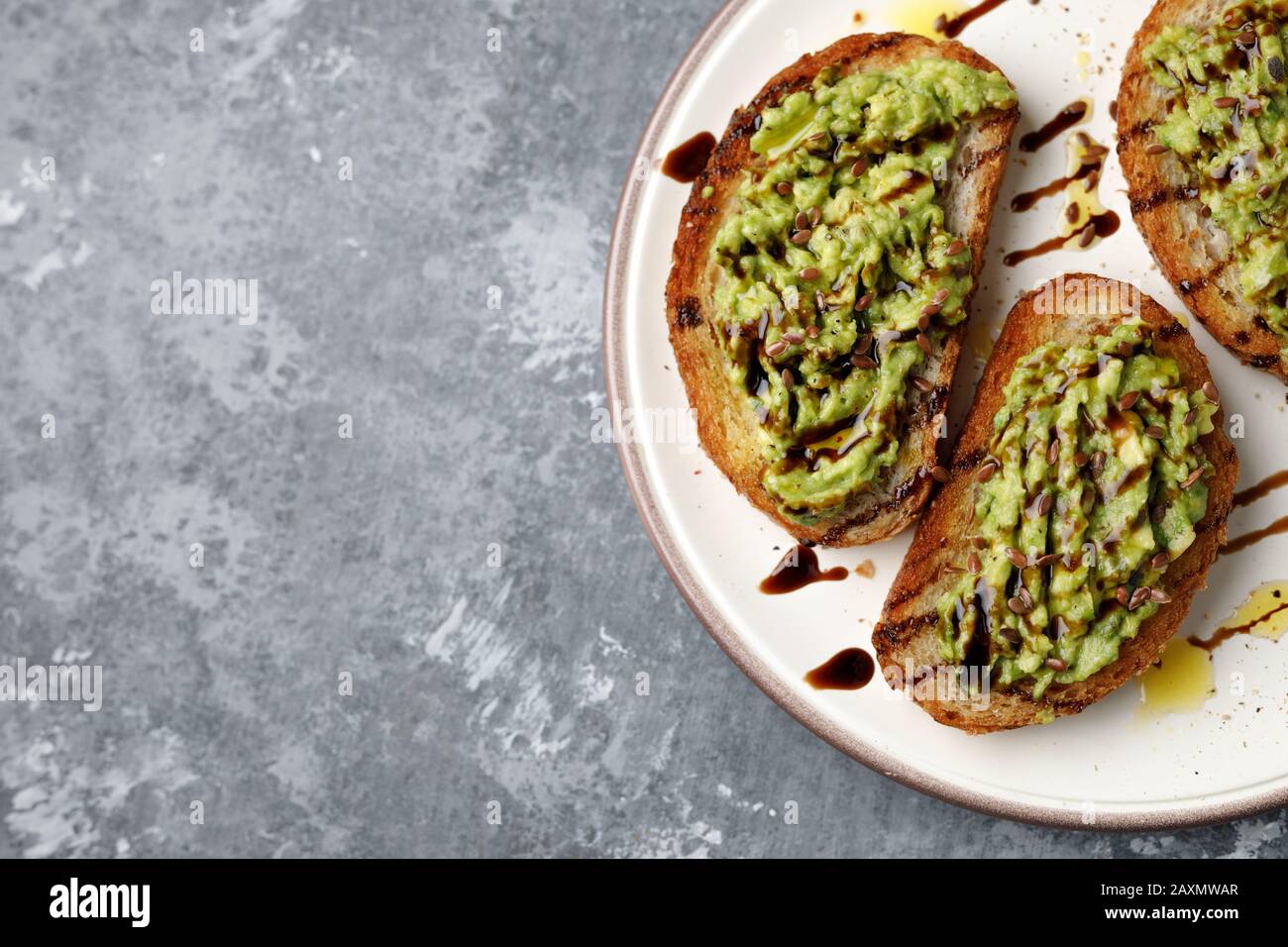 Piatto di brindisi di avocado tritato di fresco con condimento di balsamico, lino o semi di lino e olio su una piastra bianca, su uno sfondo grigio Foto Stock