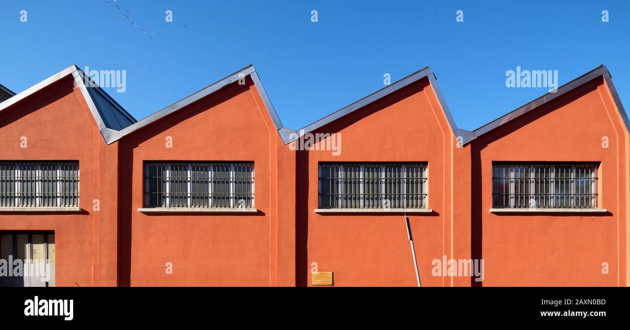 Capannone di fabbrica vecchio stile con una profonda facciata arancione su un cielo blu, tetto a zig-zag e finestre nella periferia meridionale di Milano, Italia Foto Stock