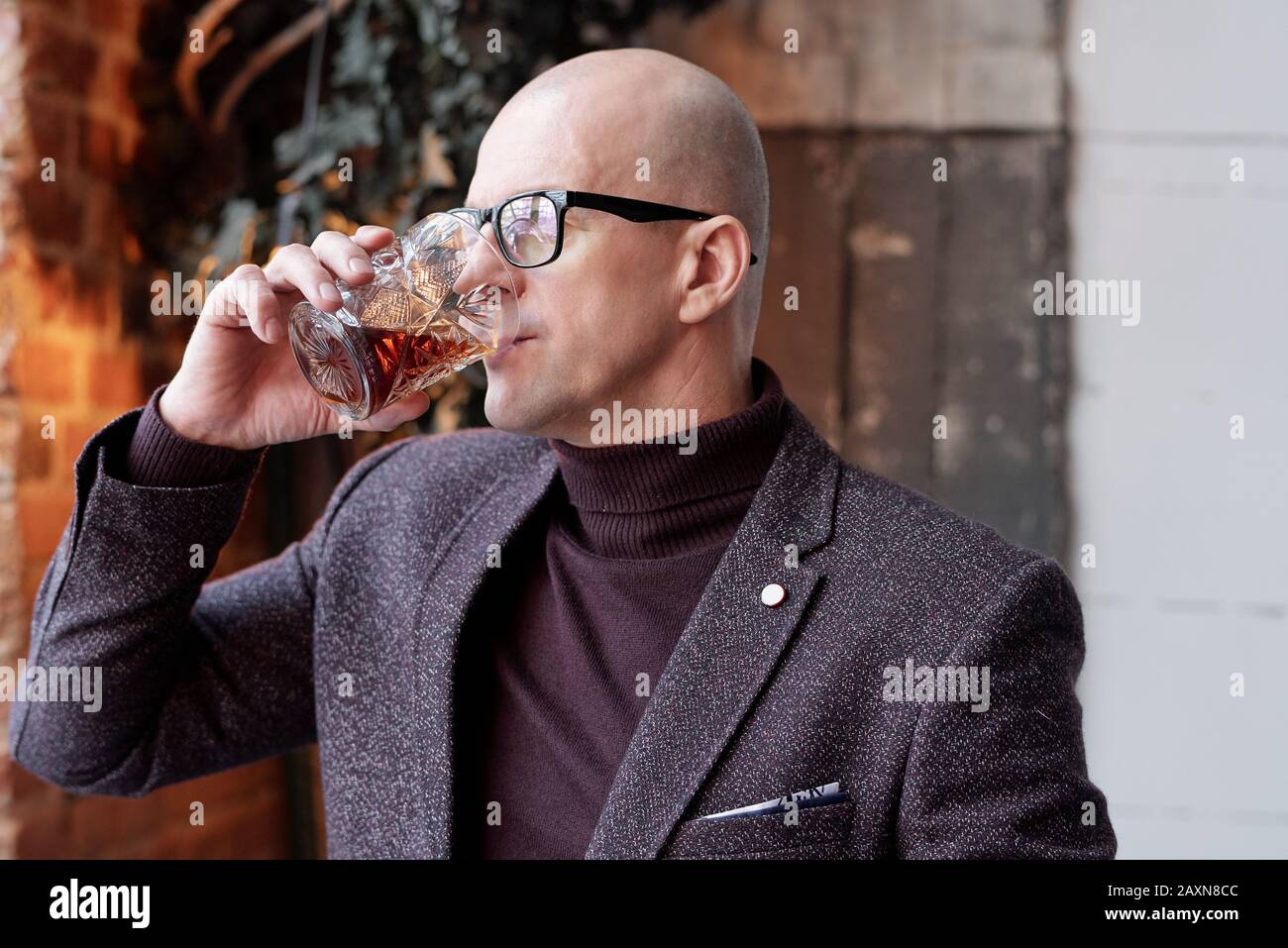 Ricco uomo calvo di mezza età in bicchieri in piedi nel ristorante loft e bere alcolici dal vetro Foto Stock