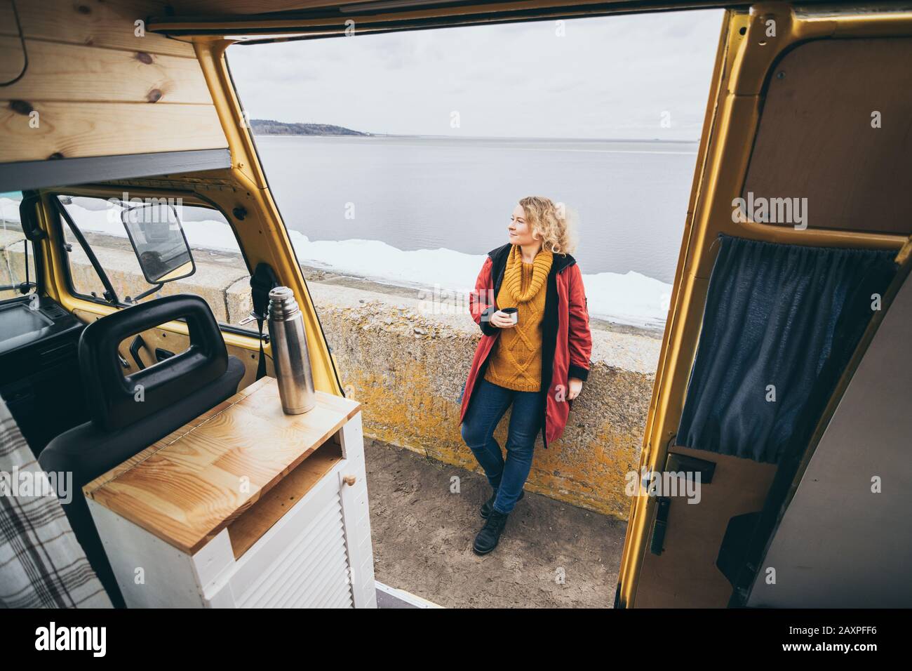 Giovane donna bionda accanto al camper che si affaccia sul mare invernale ghiacciato. Vista attraverso lo sportello aperto. Foto Stock