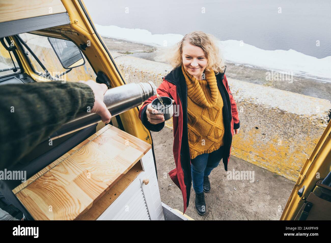 Giovane donna bionda accanto al camper che si affaccia sul mare invernale ghiacciato. Vista attraverso lo sportello aperto. Foto Stock
