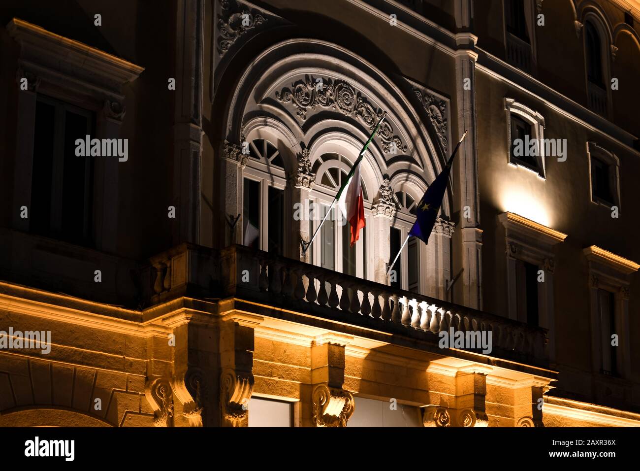 La facciata di un palazzo del governo italiano di notte con il balcone illuminato che mostra le bandiere italiane ed europee dell'Unione a Brindisi Italia. Foto Stock