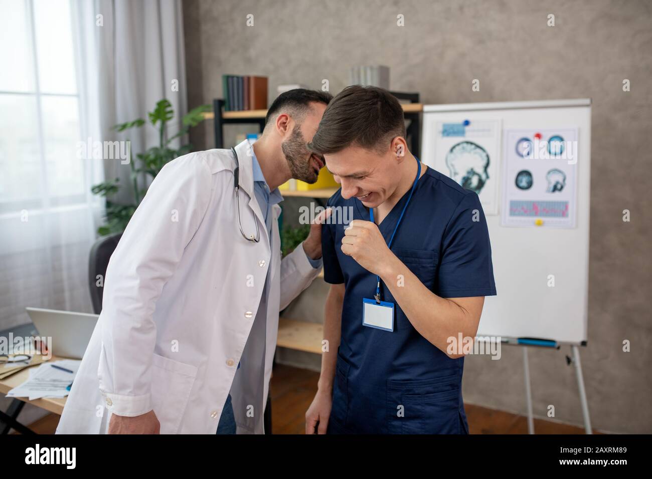 Bearded medico che dice qualcosa di divertente onn l'orecchio al suo collega Foto Stock