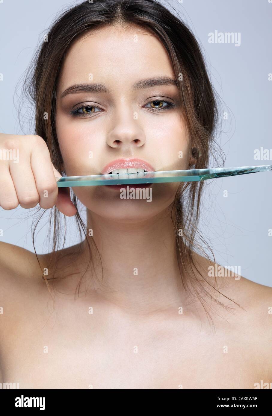 Ritratto di una bella ragazza che mordente uno specchio splinter. Femmina con specchio in mano che si posa su sfondo grigio. Foto Stock