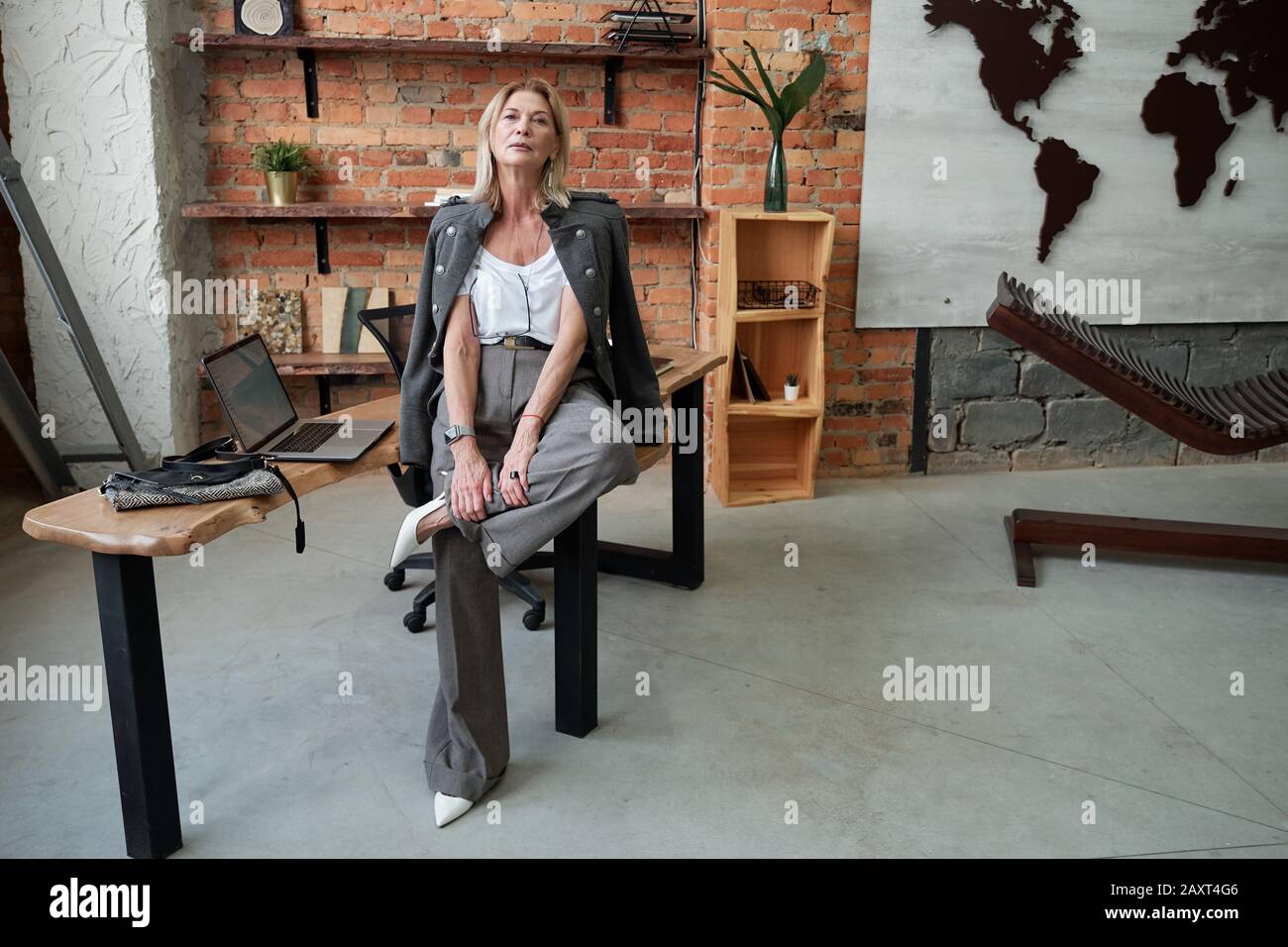 Donna matura e moderna e sicura in un abito elegante seduto sul tavolo e in posa per la fotocamera in ufficio loft Foto Stock
