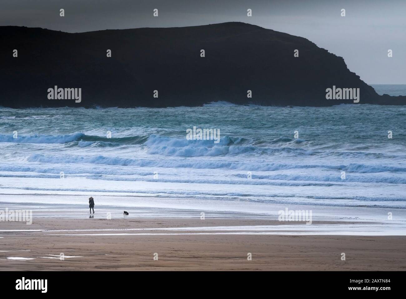 Una vista distante di un camminatore del cane solitario e del suo cane da compagnia che cammina lungo la riva in una gelida giornata sulla spiaggia di Fistral a Newquay in Cornovaglia. Foto Stock