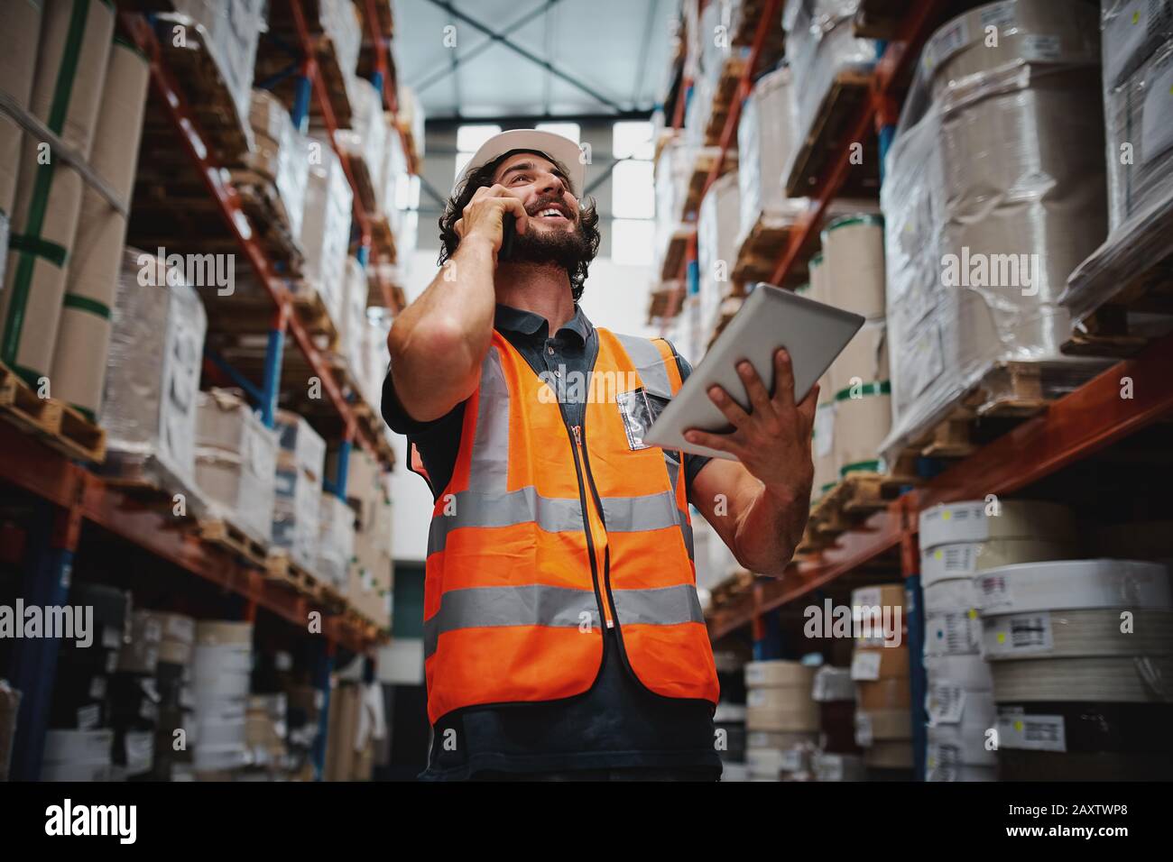 Vista ad angolo basso del responsabile del magazzino che parla al telefono mentre sorride e tiene il tablet digitale in piedi nella corsia con le merci Foto Stock