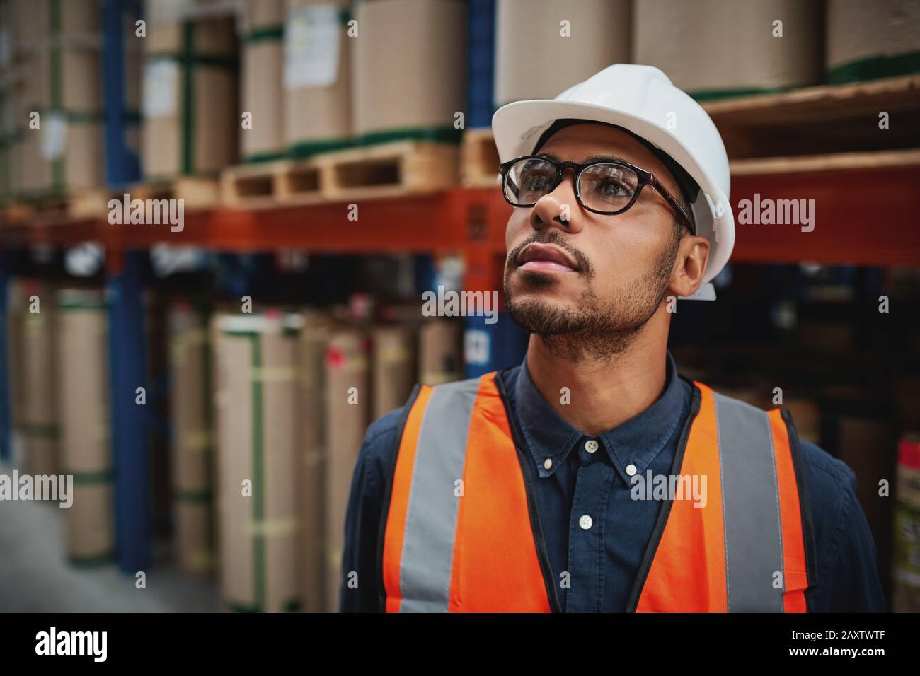 Supervisore maschile di magazzino in uniforme e il pensiero casco in piedi in fabbrica guardando lontano Foto Stock