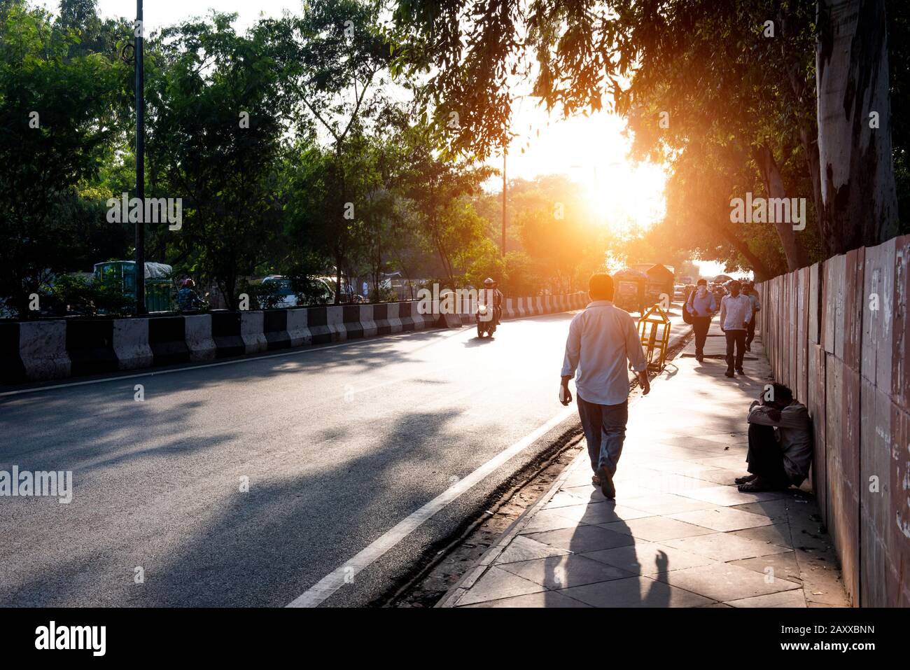 Le persone che camminano per lavorare subito dopo l'alba lungo una trafficata strada di Nuova Delhi con luce del sole e un mendicante sul marciapiede Foto Stock