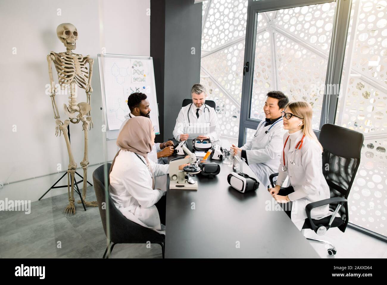 Team multietnico di giovani medici scienziati che hanno conversato, preparando a un nuovo progetto scientifico. Moderna sala da laboratorio, scheletro umano e portadocumenti Foto Stock
