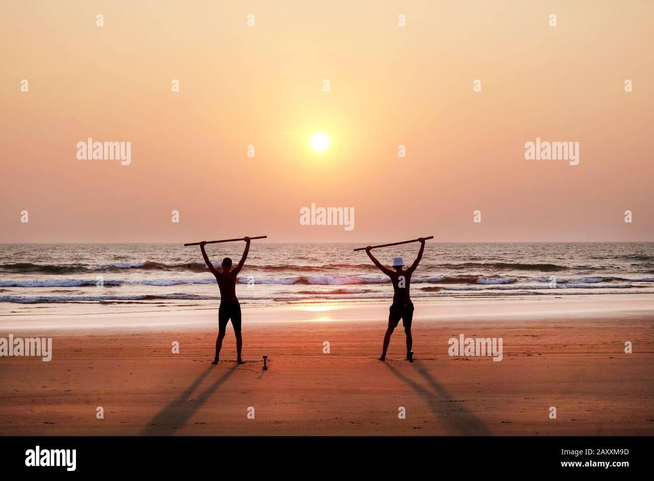 due uomini irriconoscibili che si esercitano al tramonto tenendo due pali di legno come pesi su un'ampia spiaggia di sabbia vuota, sono in piedi con le braccia Foto Stock