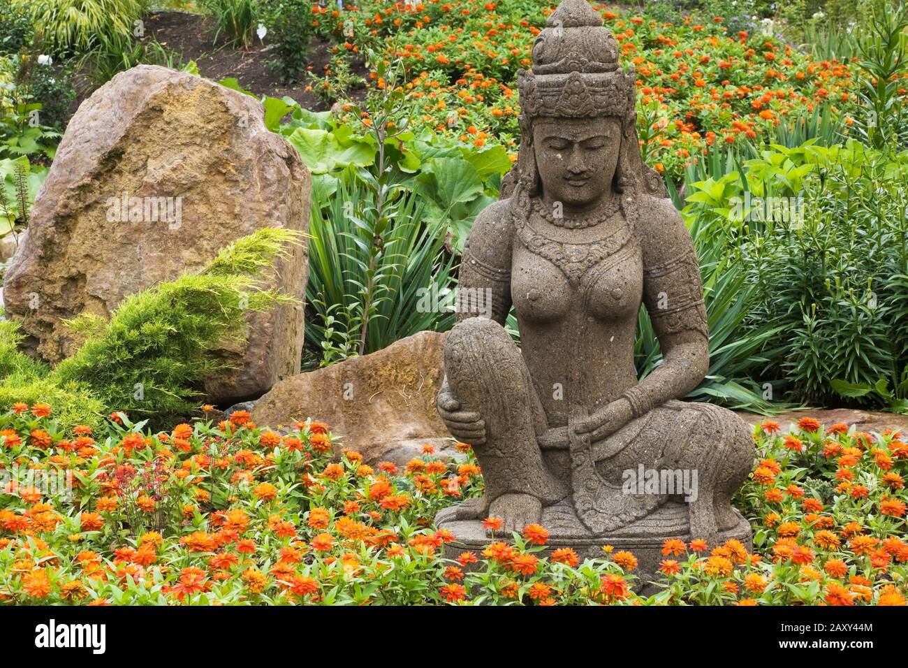 Pietra marrone scultura dea indonesiana in arancio Zinnia fiore letto nel giardino Zen in estate al giardino Route des Gerbes d'Angelica. Foto Stock