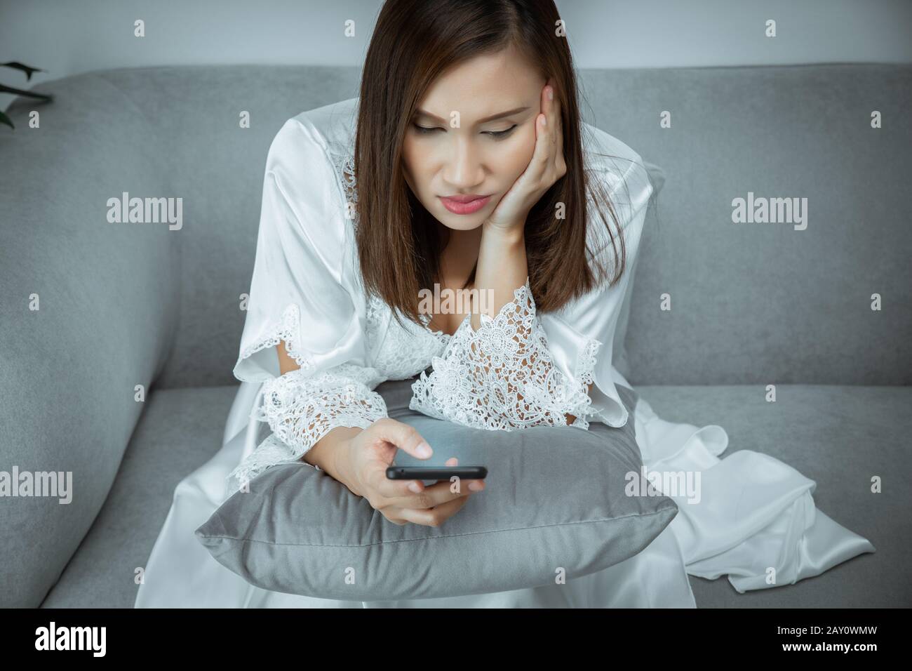 Donna che indossa laccio da notte e lungo bianco limo uso del telefono cellulare e seduta sul divano grigio di notte, concetto Mobile addict, non può dormire Foto Stock