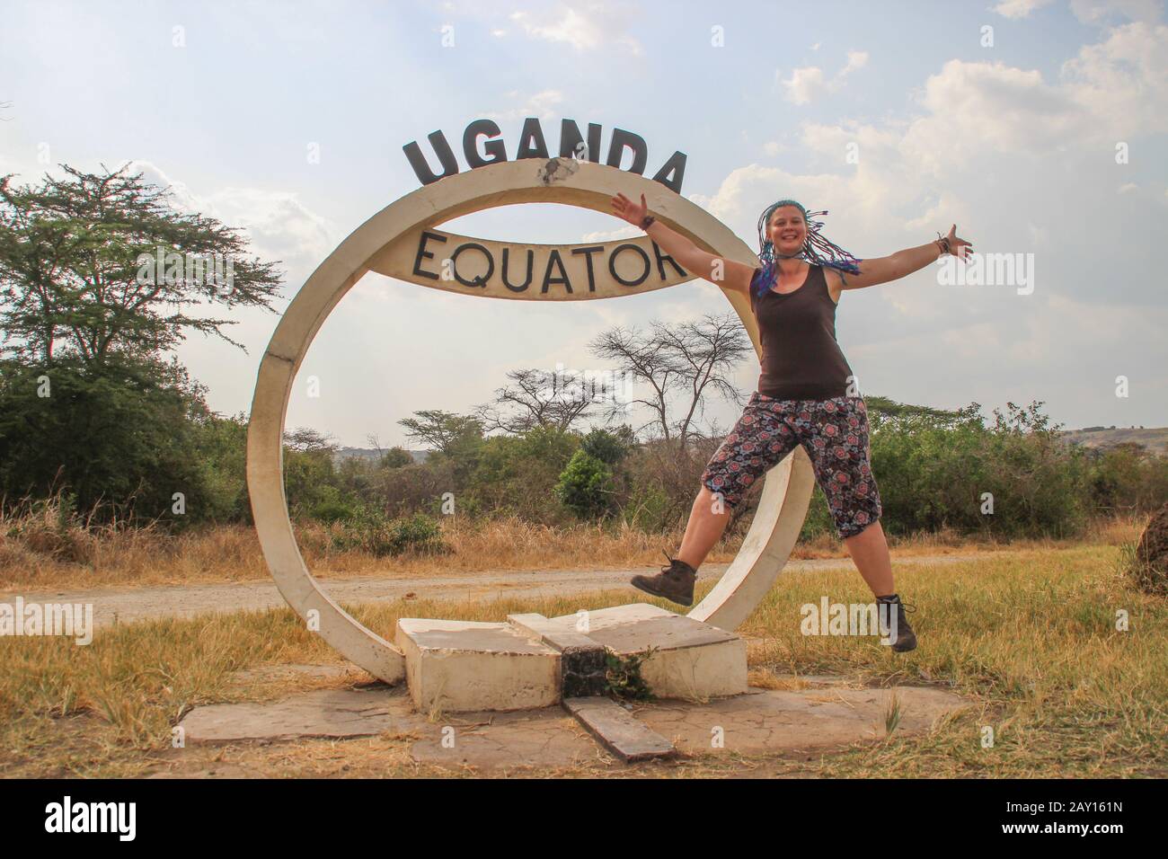 North-South Boundary Mark - Equatore nel Parco Nazionale della Regina Elisabetta in Uganda e una gioiosa ragazza turistica europea caucasica che salta. Foto Stock