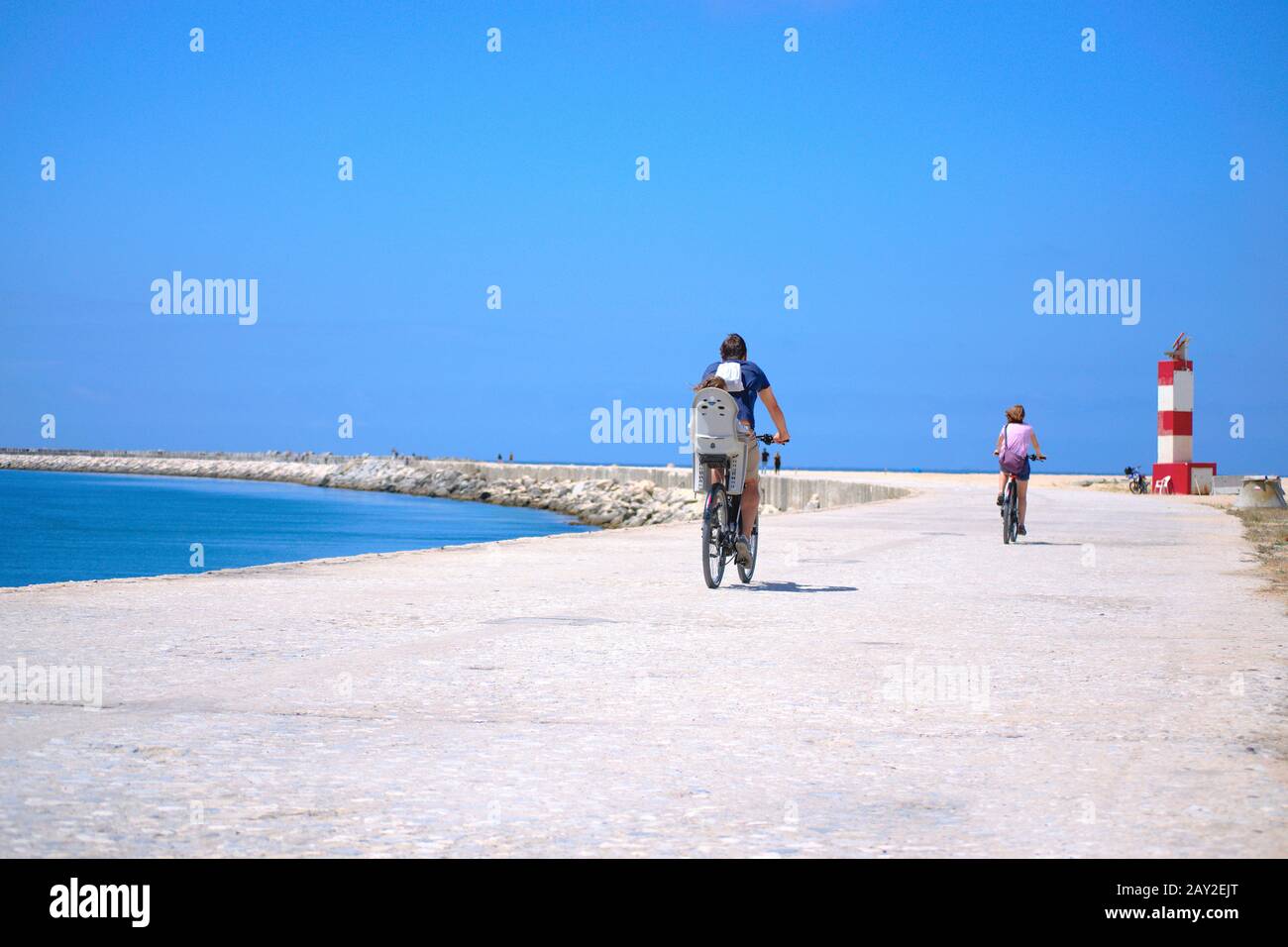 Coppia e il loro bambino che guida biciclette verso una piccola casa leggera in una calda giornata di estati accanto a una bella spiaggia blu. Foto Stock
