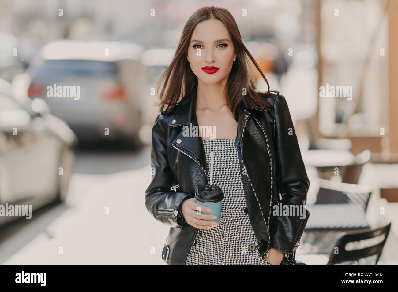 Bella donna alla moda con trucco, labbra dipinte di rosso chiaro, indossa giacca in pelle nera, passeggini in ambiente urbano, stand vicino alla caffetteria, tiene prendere Foto Stock