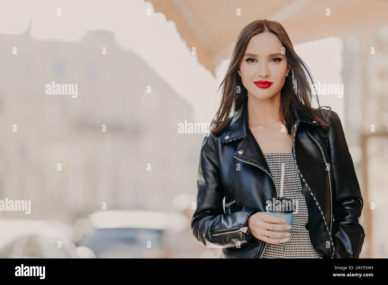 Un tocco all'aperto di un modello femminile millenario con trucco, vestito con elegante giacca in pelle, bevande a base di caffè da tazza usa e getta, girovaga intorno alla c. Foto Stock