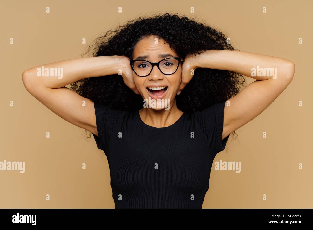 Giovane donna con capelli ricci afro copre le orecchie, grida come le esigenze essere tranquilla, cant stand rumore, mantiene la bocca aperta, indossa occhiali e casual nero t sh Foto Stock