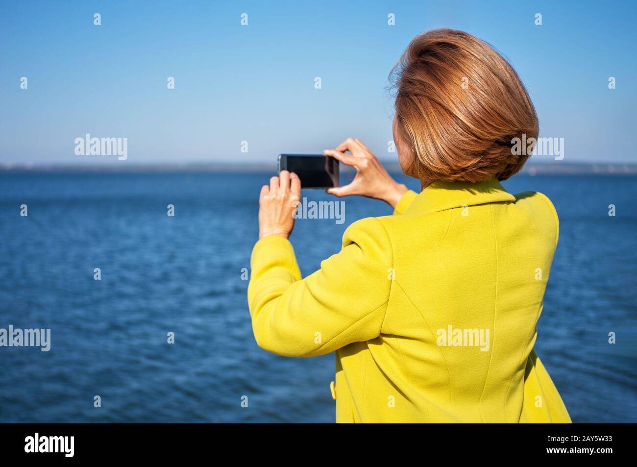 Elegante giovane donna in un cappotto giallo brillante scatta foto del mare utilizzando uno smartphone. Telefono cellulare a schermo vuoto. Primo piano. Copia fase. Foto Stock