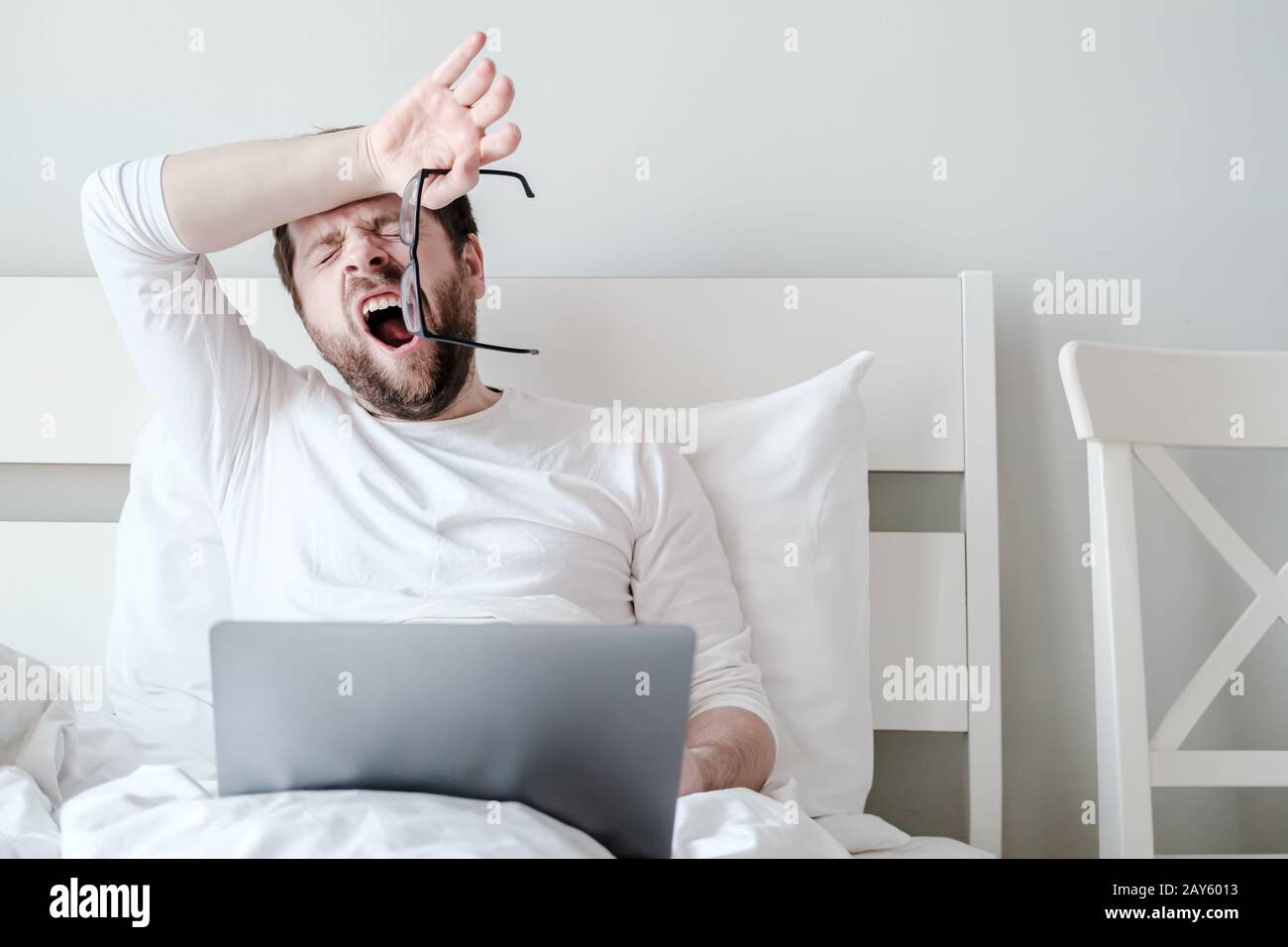 L'uomo tolse occhiali e sveglie, non ha avuto abbastanza sonno, funziona con un portatile in un letto morbido, a casa, al mattino presto. Foto Stock