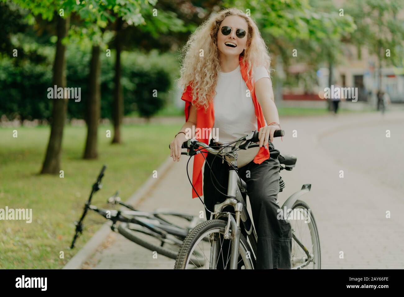 Piacevole giovane donna in occhiali da sole, passeggiate in bicicletta sportiva, soste per la pausa, gode di bellezza della natura, atmosfera calma, ride positivamente, mantiene b Foto Stock