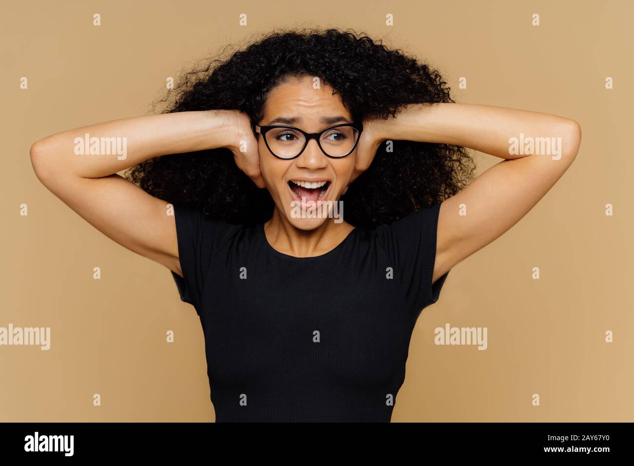 L'immagine della donna afroamericana impressionabile copre le orecchie, urla ad alta voce, richieste di rumore di cant stand spegnere la musica, vestito in t shirt nero casual, ottico Foto Stock